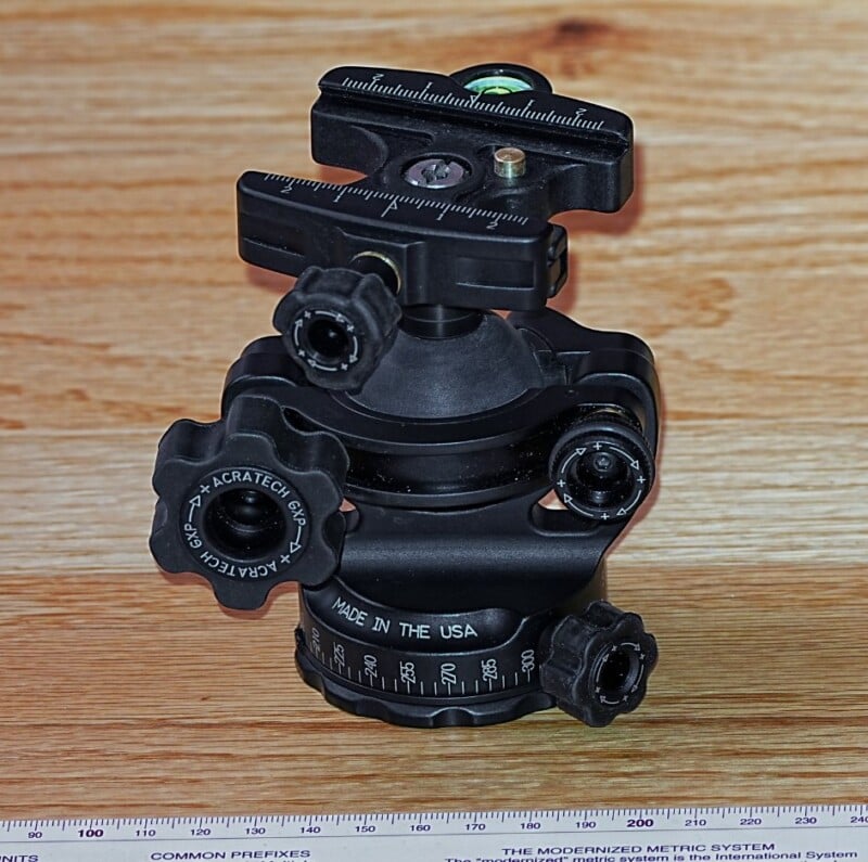 A black Acratech GP-SS ball head with adjustment knobs and a built-in bubble level sits on a wooden surface. A ruler is placed next to it for size reference.