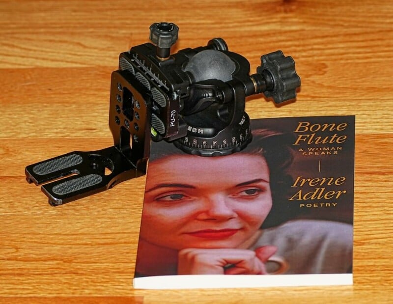 A black mechanical device sits on a wooden table next to a book titled "Bone Flute: A Woman Speaks" by Irene Adler. The book cover features a portrait of a woman with her hand on her chin.