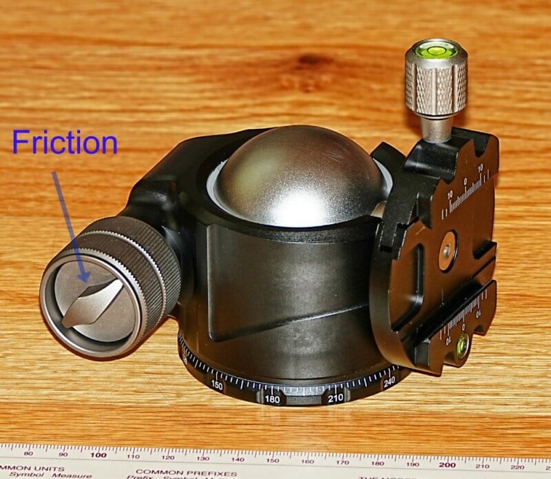 A black and silver panoramic tripod head on a wooden surface. It features friction control, a leveling bubble, and an angle scale. The word "Friction" is labeled in blue near the dial. A ruler is partially visible at the base.