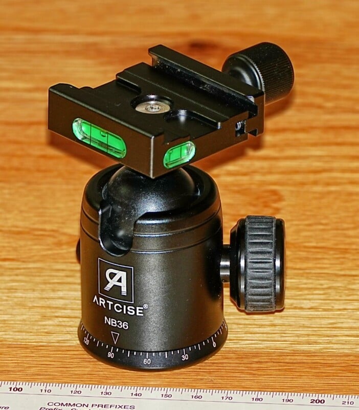A black Arca-Swiss style ball head, labeled ARTCTISE NB36, is placed on a wooden surface. It has two green bubble levels and adjustment knobs. A ruler marked in centimeters and inches is positioned in the foreground.