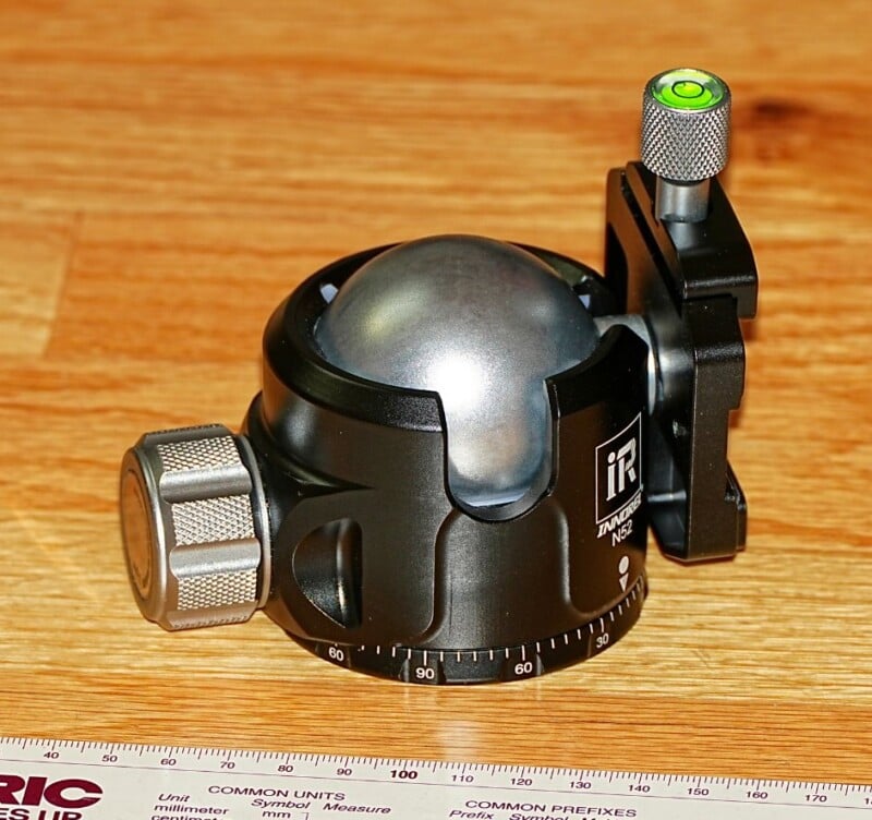 A black and silver camera tripod ball head sits on a wooden surface, featuring adjustment knobs and a visible bubble level. A ruler with measurement markings is partially visible in the foreground.