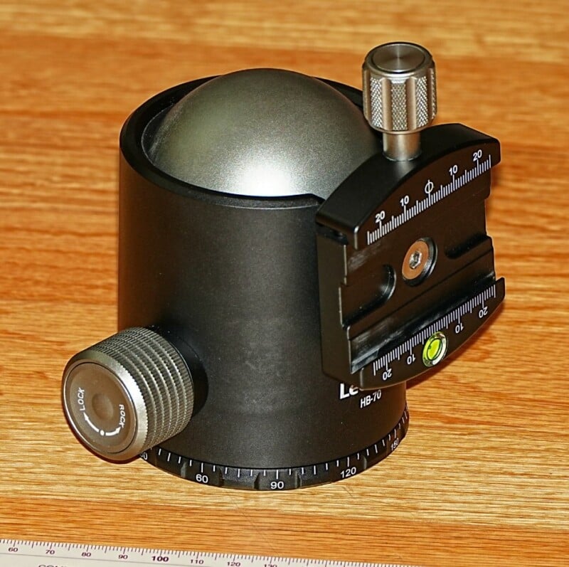 A black tripod head with a spherical design, featuring adjustment knobs and a prominent vertical grip. It has measurement markings and a built-in level, all set on a wooden surface.