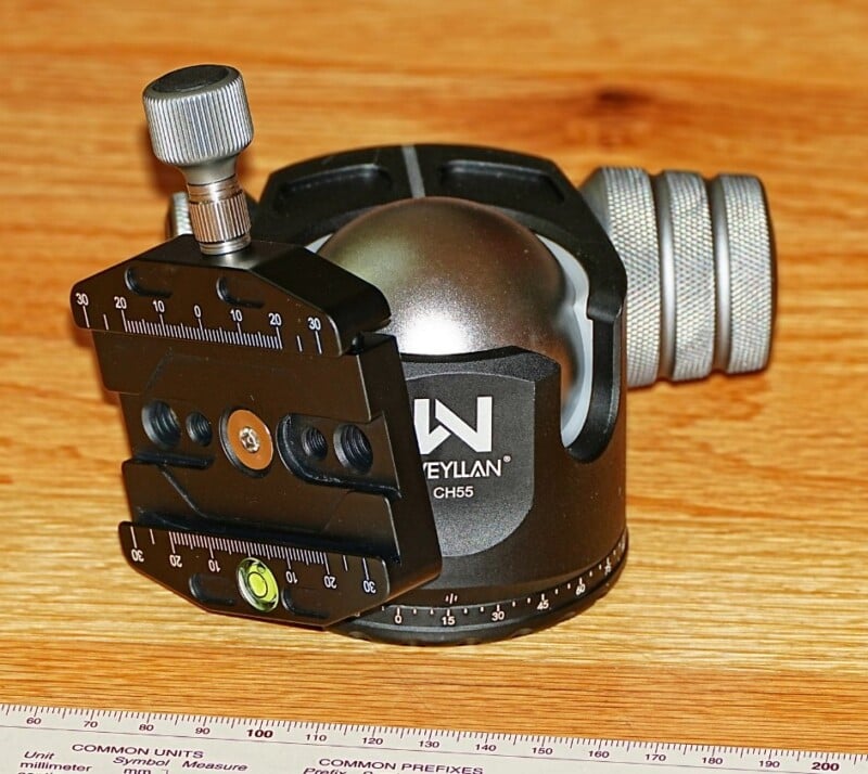 A black and silver camera tripod head with adjustment knobs is sitting on a wooden surface. It features measurement markings and has a bubble level indicator. A ruler with unit conversions is partially visible at the bottom.