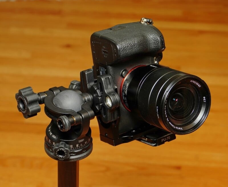 A black camera with a large lens is mounted on a tripod with a ball head. The background features a wooden floor, giving the image a warm tone.