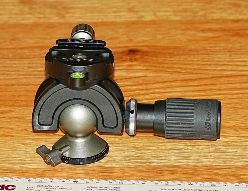 A close-up of a black and gray tripod head with adjustment knobs and a spirit level rests on a wooden surface. A printed scale is partially visible along the bottom edge.