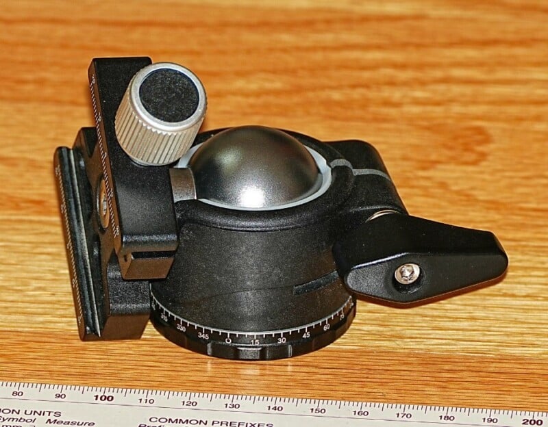 A black ball head tripod mount with a metallic knob and adjustment lever is placed on a wooden surface. A measuring scale is positioned in front of it, displaying units in millimeters.