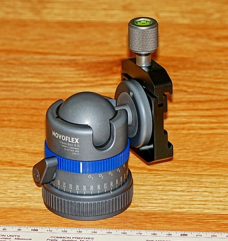Ball head camera mount with a blue ring and a gray knob is placed on a wooden surface. It has a leveling bubble on top and is next to a ruler with measurement units.
