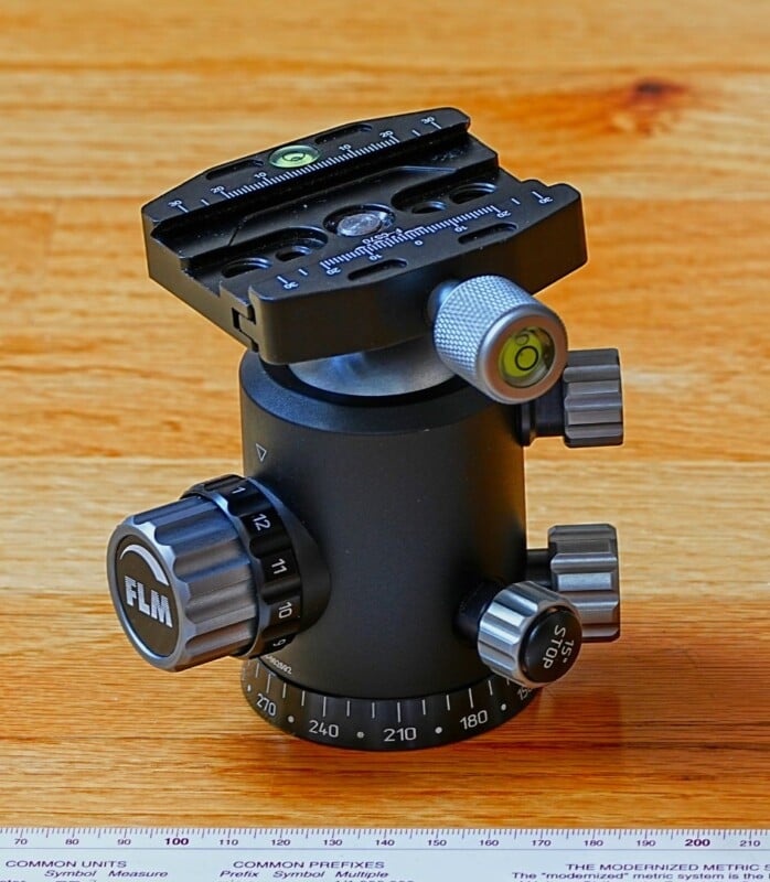 A black metal tripod head with various adjustment knobs and a leveling bubble sits on a wooden surface. The base has degree markings. A ruler is partially visible at the bottom, showing common units.