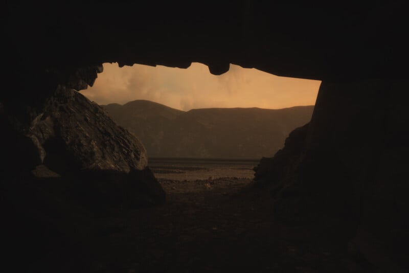 View from inside a dark cave looking out towards a landscape with distant mountains under a hazy, orange sky. The rugged cave opening frames the scene, casting shadows on the rocky surface inside.
