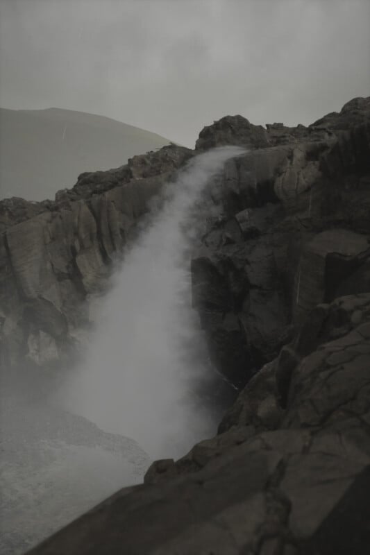 A dramatic, misty waterfall cascades down from jagged, dark rocks under a cloudy, overcast sky. The forceful water creates a mist that obscures part of the rock face, adding an ethereal atmosphere to the scene.