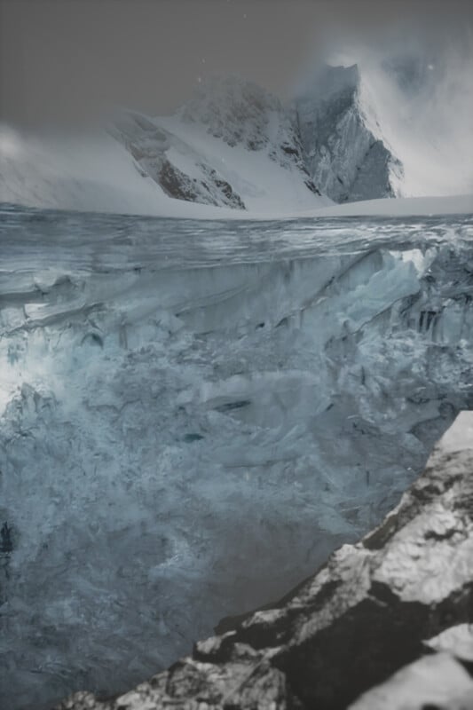A vast, rugged glacier with deep blue ice and jagged crevices in the foreground. Snow-covered mountains loom in the background under a cloudy sky, creating a dramatic and cold landscape.