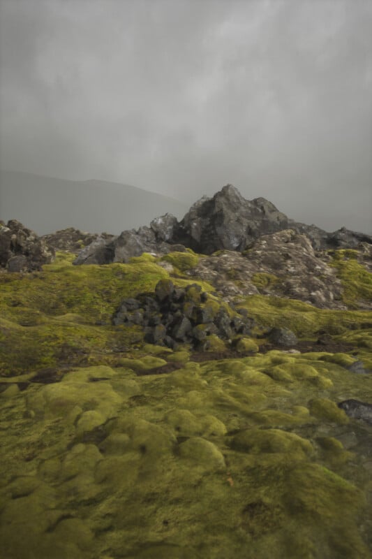 A landscape of moss-covered rocks under a gray, overcast sky. The scenery is slightly foggy, lending a mystical appearance. The terrain is uneven, with clusters of larger rocks scattered across the mossy ground.