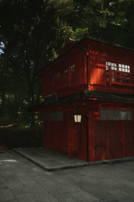 A traditional Japanese building with red wooden walls and a gray tiled roof is nestled among lush green trees. The sunlight filters through the foliage, casting patterns on the building and stone pathway in front.