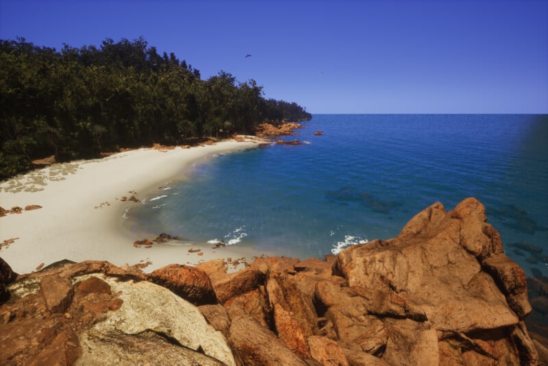 A scenic view of a secluded beach with white sand and clear blue water. Rocky formations in the foreground lead to a bay surrounded by dense, green forest. A clear blue sky stretches overhead with a few birds flying near the horizon.
