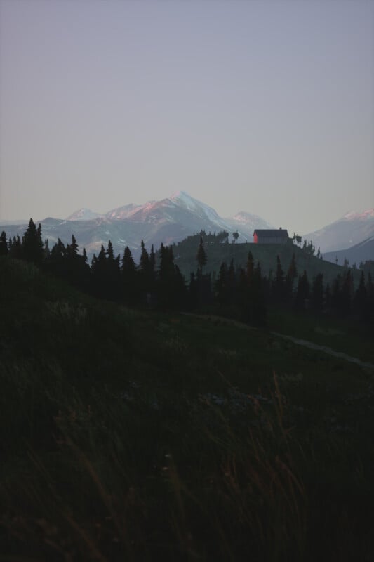 A tranquil mountain landscape at dusk with a distant cabin on a hill. Snow-capped peaks are visible against a serene sky. Tall evergreen trees dot the hillside, creating a peaceful, picturesque scene.