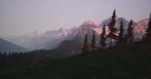 Sunset view of a mountain landscape with snow-capped peaks, tall evergreen trees, a grassy hillside, and a rustic building in the foreground. The sky is clear with a soft, pinkish glow.