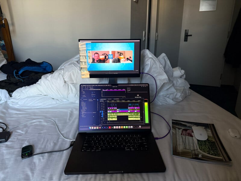 A laptop and an external monitor are set up on an unmade bed. The laptop displays video editing software, while the monitor shows a video call with several people. A magazine and a white mouse are on the bed nearby. A door is visible in the background.