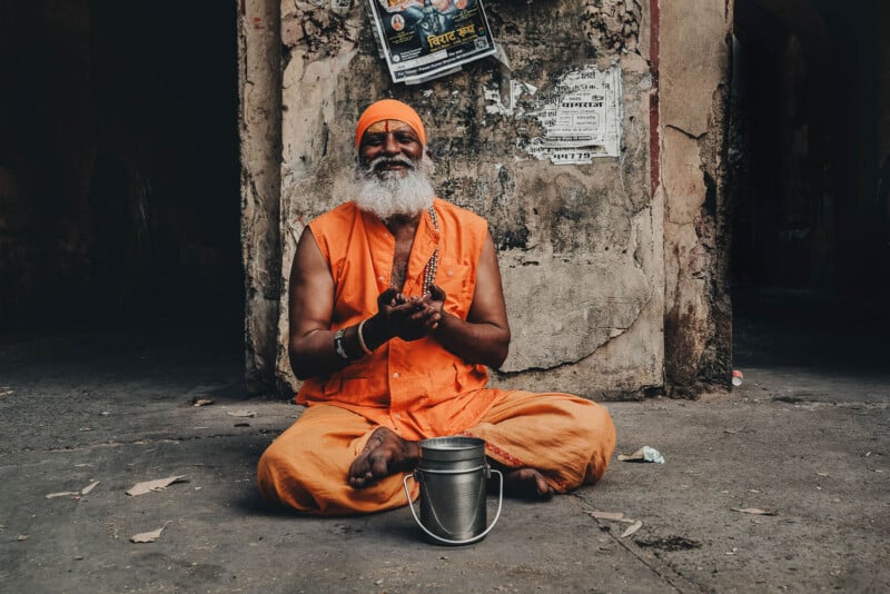 An elderly man with a white beard, dressed in bright orange robes and headscarf, sits cross-legged on a street with a small metal container in front of him, against a worn, peeling wall with posters.