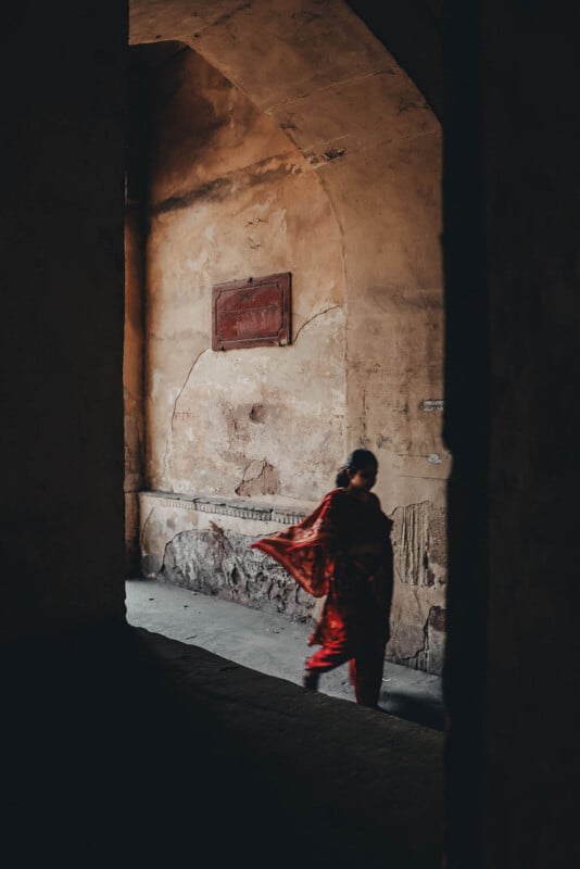 A person in a red patterned garment walks through a dimly lit, weathered archway with cracked beige walls and a faded sign above. The figure appears blurry, emphasizing motion.