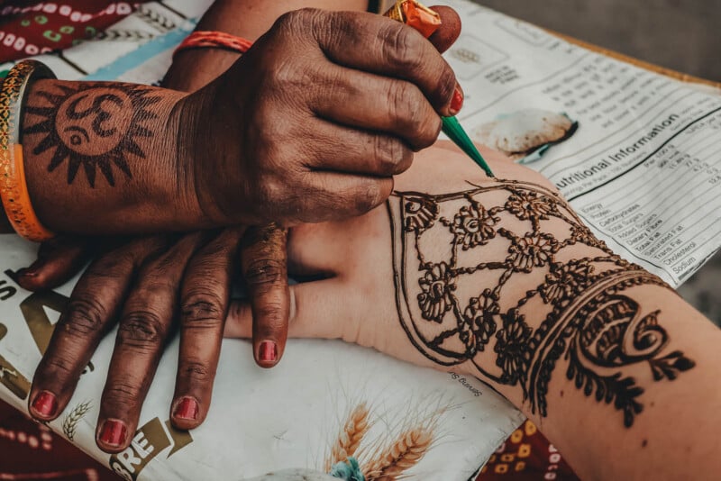 A person applies intricate henna designs on another person’s hand, while the artist's own hand shows a simple sun-shaped henna tattoo. Both hands rest on a newspaper.