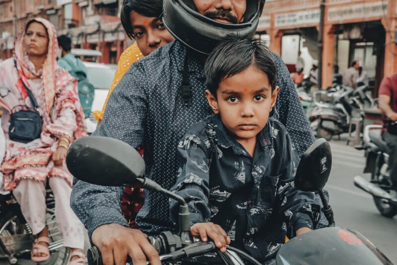 A young boy sits in front of an adult on a motorcycle in a busy street. The boy looks directly at the camera with a serious expression. People and parked motorcycles are visible in the background.