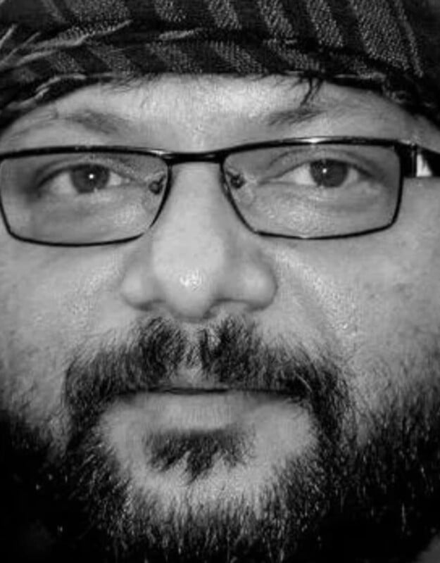 Black and white close-up of a bearded person wearing rectangular glasses and a patterned headscarf, looking slightly past the camera.