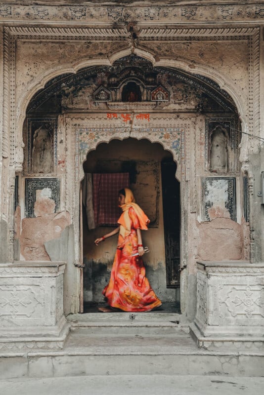 A woman in a vibrant orange sari walks past the entrance of an ornate, weathered stone building adorned with intricate carvings and faded artwork.