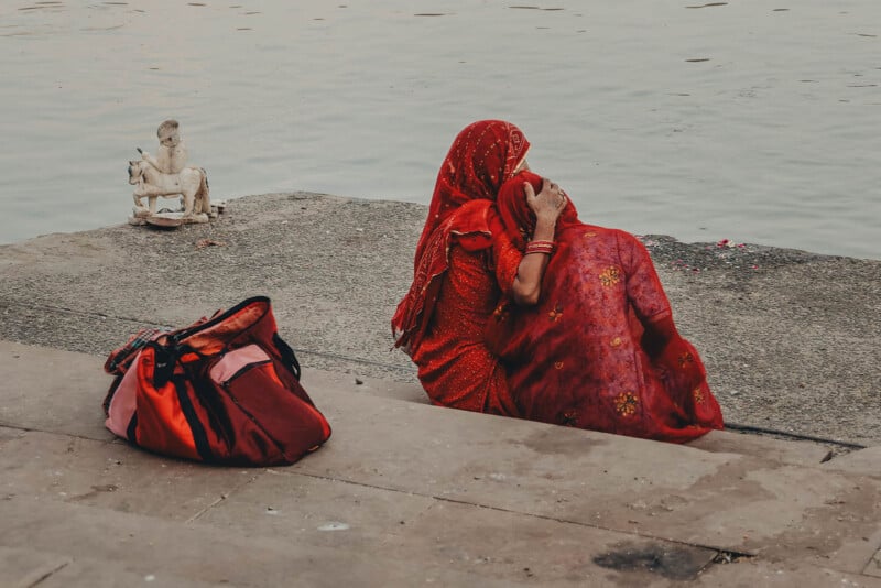A woman in a red sari sits beside a young child by a riverbank, both facing the water. A red bag lies nearby on the steps, and a small statue is visible in the background near the river.