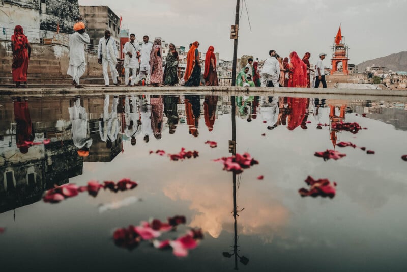 A group of people in colorful traditional clothing walk beside a large puddle reflecting their images, scattered with pink flower petals. Buildings and a red temple appear in the background under a cloudy sky.