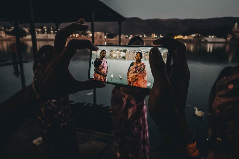 A person holds a phone, taking a picture of two women in colorful sarees by a lakeside at dusk; the women appear brightly lit on the phone screen, while the background is dark and serene.