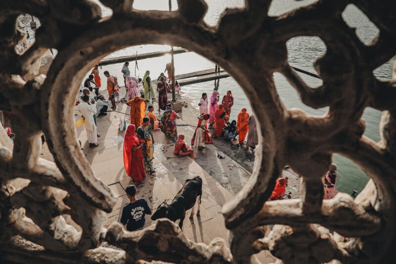 A group of people in colorful clothing gather by a riverside, viewed through an ornate stone lattice. Some are standing, some are sitting, and a cow is present near the water’s edge, with sunlight reflecting on the river.
