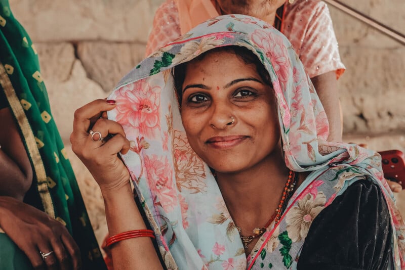 A woman wearing a floral headscarf smiles gently at the camera, holding part of the scarf with her hand. She is adorned with traditional jewelry and has a bindi on her forehead. Other people are partially visible in the background.