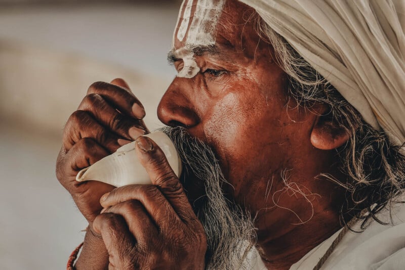 A man with white markings on his forehead and a turban blows a conch shell, holding it with both hands; his expression appears focused and serene.