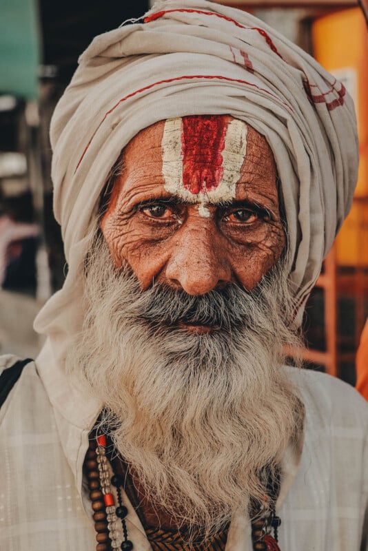 An elderly man with a long white beard and mustache, wearing a white turban and robe, stares directly at the camera. He has traditional religious markings on his forehead and beaded necklaces around his neck.