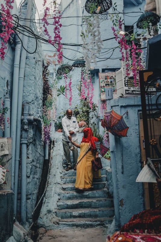 A man and woman in traditional Indian clothing walk down blue-painted steps in a narrow alley decorated with hanging flowers and umbrellas. The woman wears a yellow sari and the man wears a white shirt and pants.