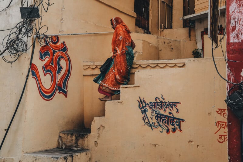 A woman in a colorful traditional sari walks up worn concrete steps beside a yellow wall with painted Hindi script and a large red Om symbol. Exposed wires hang overhead in the narrow passage.