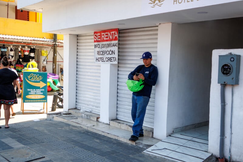 A man in a blue shirt and jeans stands leaning against a white building with a closed metal shutter, holding a green helmet and looking at his phone. A "Se Renta" sign is posted above him. People and shops are visible nearby.