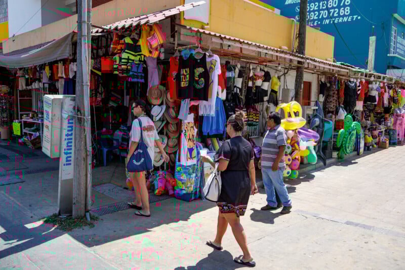 People walk past a colorful street shop selling clothes, hats, and inflatable pool toys. The shop has items hanging outside and is located on a sunny sidewalk with a telephone pole and signs nearby.