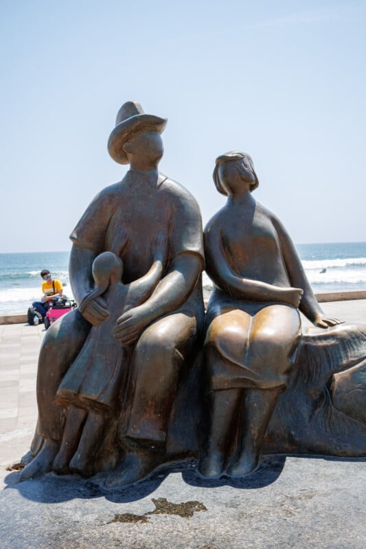 A bronze statue of a family—an adult man, woman, and child—sits together facing the sea on a seaside promenade, with waves and blue sky in the background.