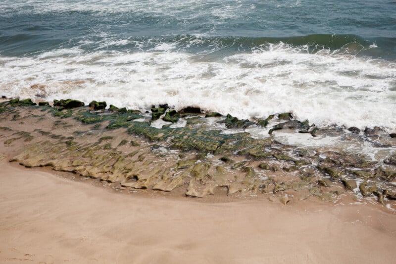 Waves wash over a sandy beach, exposing a line of flat, jagged rocks covered in green algae near the shoreline. The water appears foamy as it meets the textured, natural rock formations.