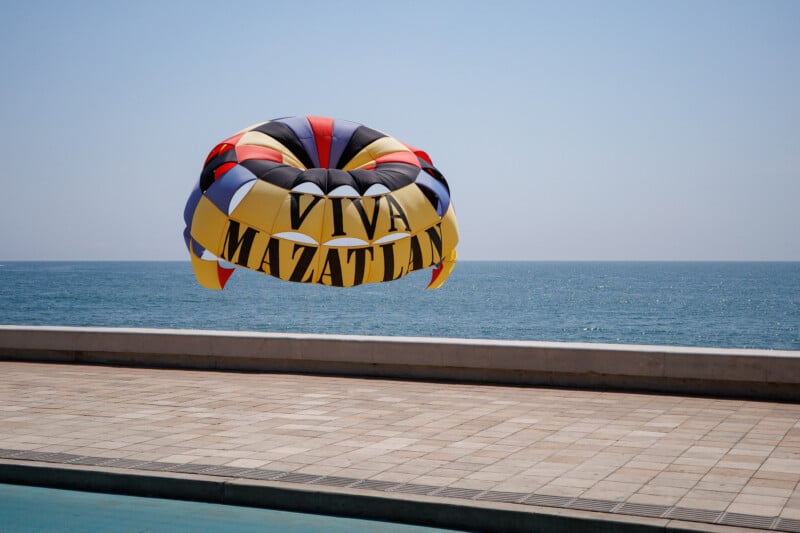 A colorful parasail with the words "Viva Mazatlán" floats above a seaside promenade, with the blue ocean and clear sky in the background.