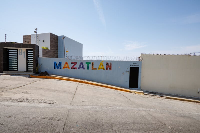 A white wall with colorful letters spelling "Mazatlán" and barbed wire on top stands next to a modern building on a sunny day. A door labeled "Privada San Martin" is visible on the right side of the wall.