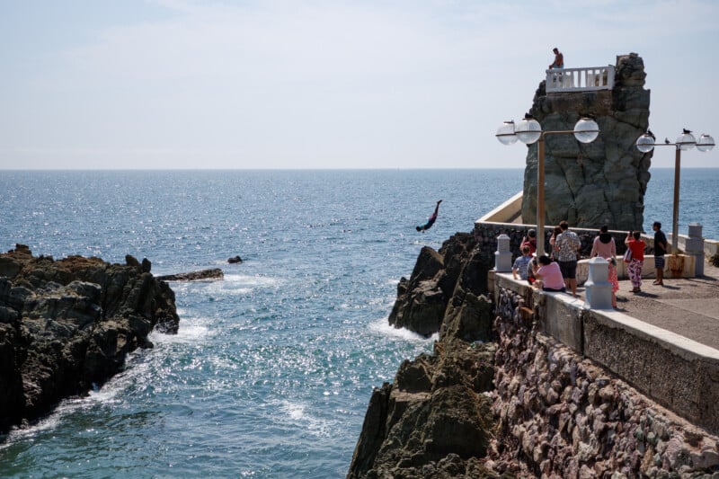 A person is diving off a rocky cliff into the ocean while a group of people watch from a nearby walkway under street lamps. The sea is calm and stretches out to the horizon under a clear sky.