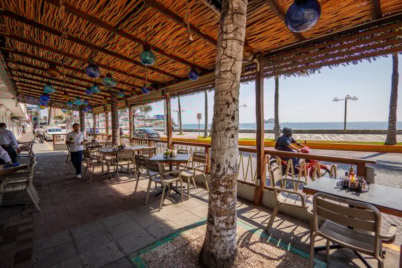 Outdoor seating area of a seaside restaurant with wooden tables and chairs under a bamboo roof, overlooking a street, palm trees, and the ocean. A person walks between tables; motorcycles and people are on the sidewalk.