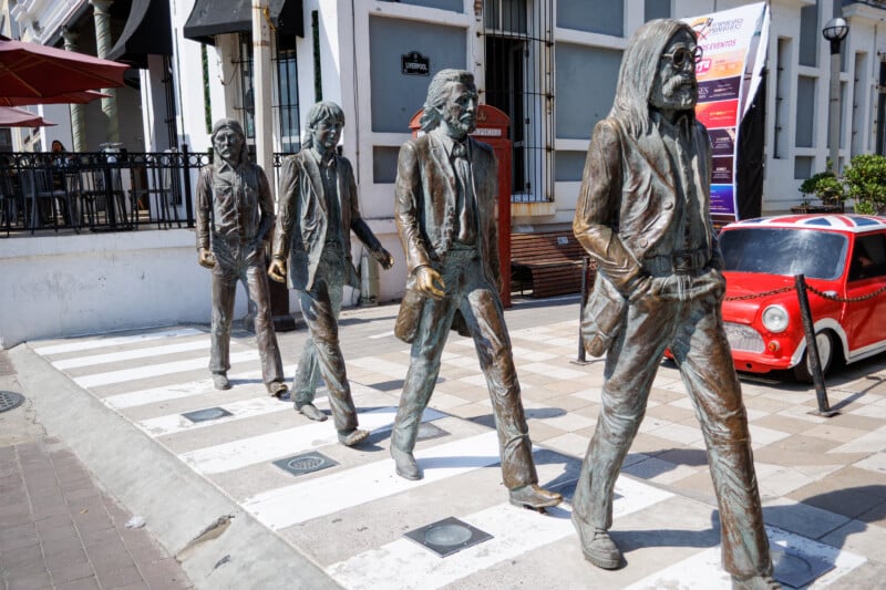 Bronze statues of four men walking across a striped crosswalk, mimicking the famous Beatles Abbey Road album cover, with buildings and a red car in the background.