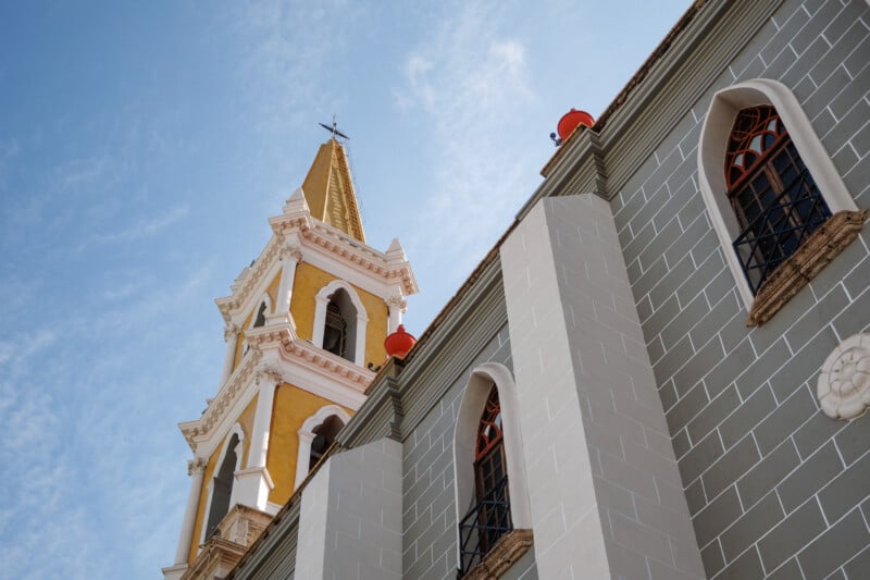 A close-up view of a church with a yellow and white bell tower, arched windows, and a pastel gray facade, set against a blue sky with wispy clouds.