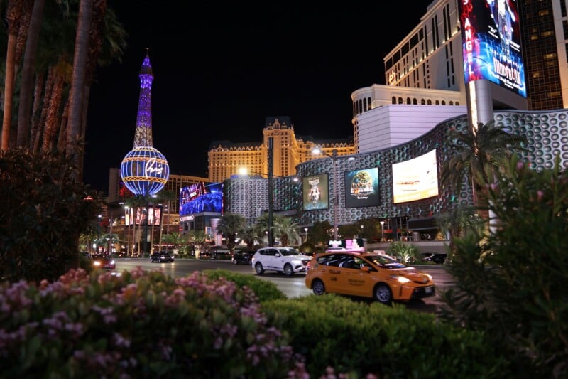 Night view of a bustling Las Vegas street with neon lights. A replica of the Eiffel Tower and a globe structure are prominent. Cars, including a yellow taxi, drive along the road, surrounded by lush greenery and brightly lit billboards.