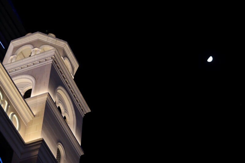 A white, illuminated clock tower against a night sky. The moon is visible in the upper right corner, partially full. The tower features arched windows and a pointed roof.