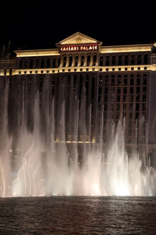 Illuminated fountains spraying water in front of the Caesars Palace hotel at night. The hotel façade is grand and classical in style, with the building name glowing in bright lights above.