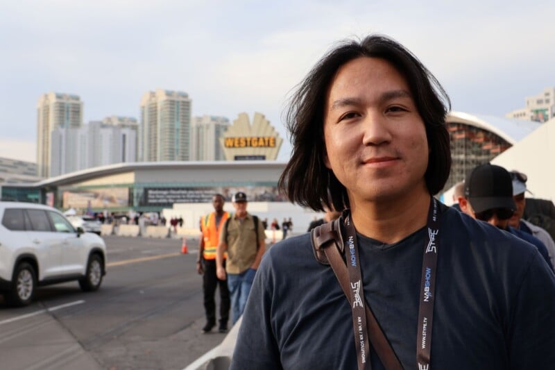 A person with long hair, wearing a black shirt and lanyard, stands outdoors. In the background, a hotel with the sign "Westgate" is visible, along with several high-rise buildings. Other people and a white SUV are also present.